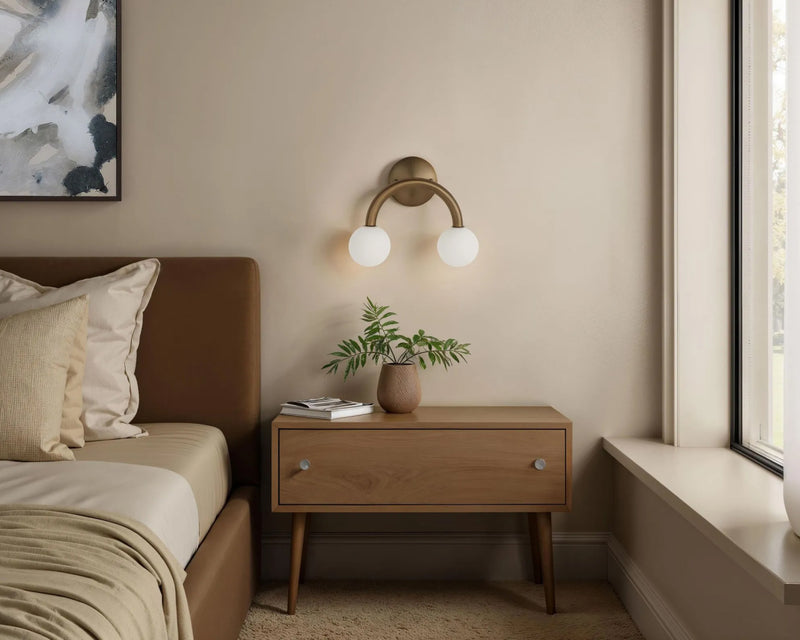 Brushed brass dual-globe arch wall sconce with frosted glass balls above oak nightstand in soft beige bedroom